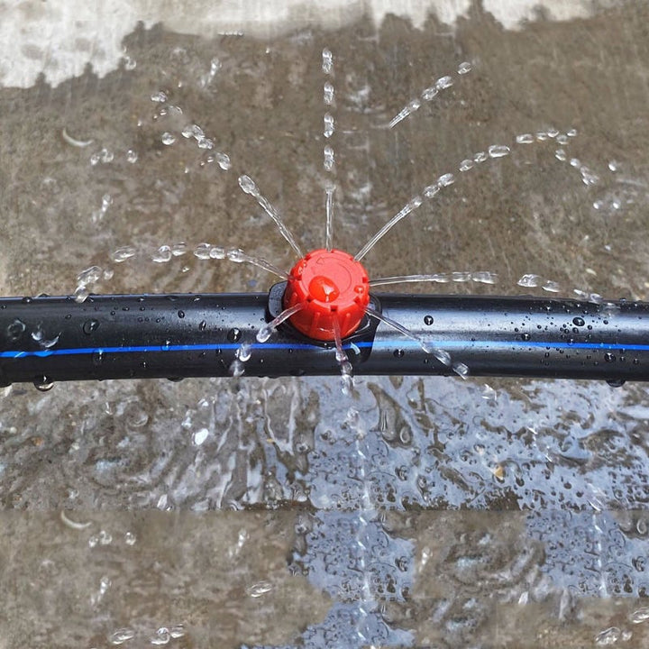 Piros csepegtetőfej a kék csíkos fekete tömlőn, ahogy vízpermetet fröcsköl a nedves felületre.
