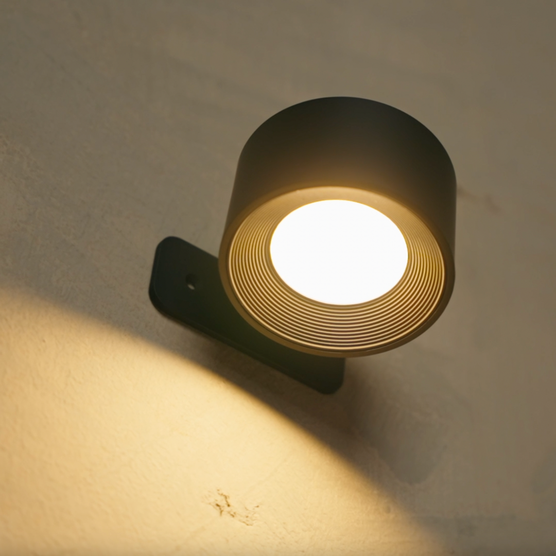 Közeli kép a fekete LED fali lámpa bekapcsolt fejéről, körkörös bordázattal és a falra vetülő fénnyel.
