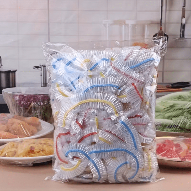 Átlátszó zacskó tele színes gumis szegélyű ételtakaró sapkákkal a konyhapulton, tálak és tányérok között.