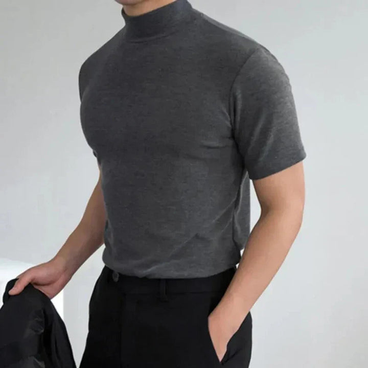 Szürke, testhez simuló magas nyakú póló modellen, fekete nadrággal, az egyik keze zsebben, a másikban kabátot tart. 3