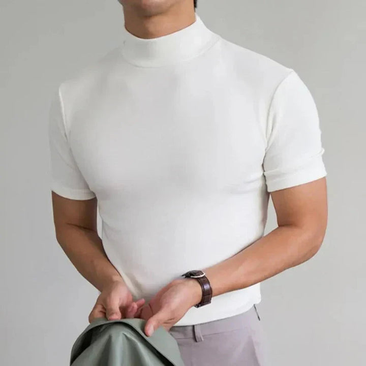 Fehér, rövid ujjú, magas nyakú póló modellen, csuklóján órával, világos nadrággal, kezében zöldesszürke kabát. 4