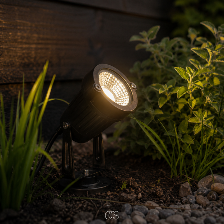 Fekete LED kerti reflektor a talajban egy sötét faburkolat mellett, fű és kavics között, közeli felvétel.