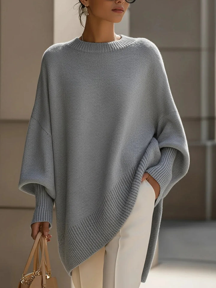 Szürke oversize pulóver modellen, elölről, bő ujjakkal, bordás szegéllyel és fehér nadrággal. 9
