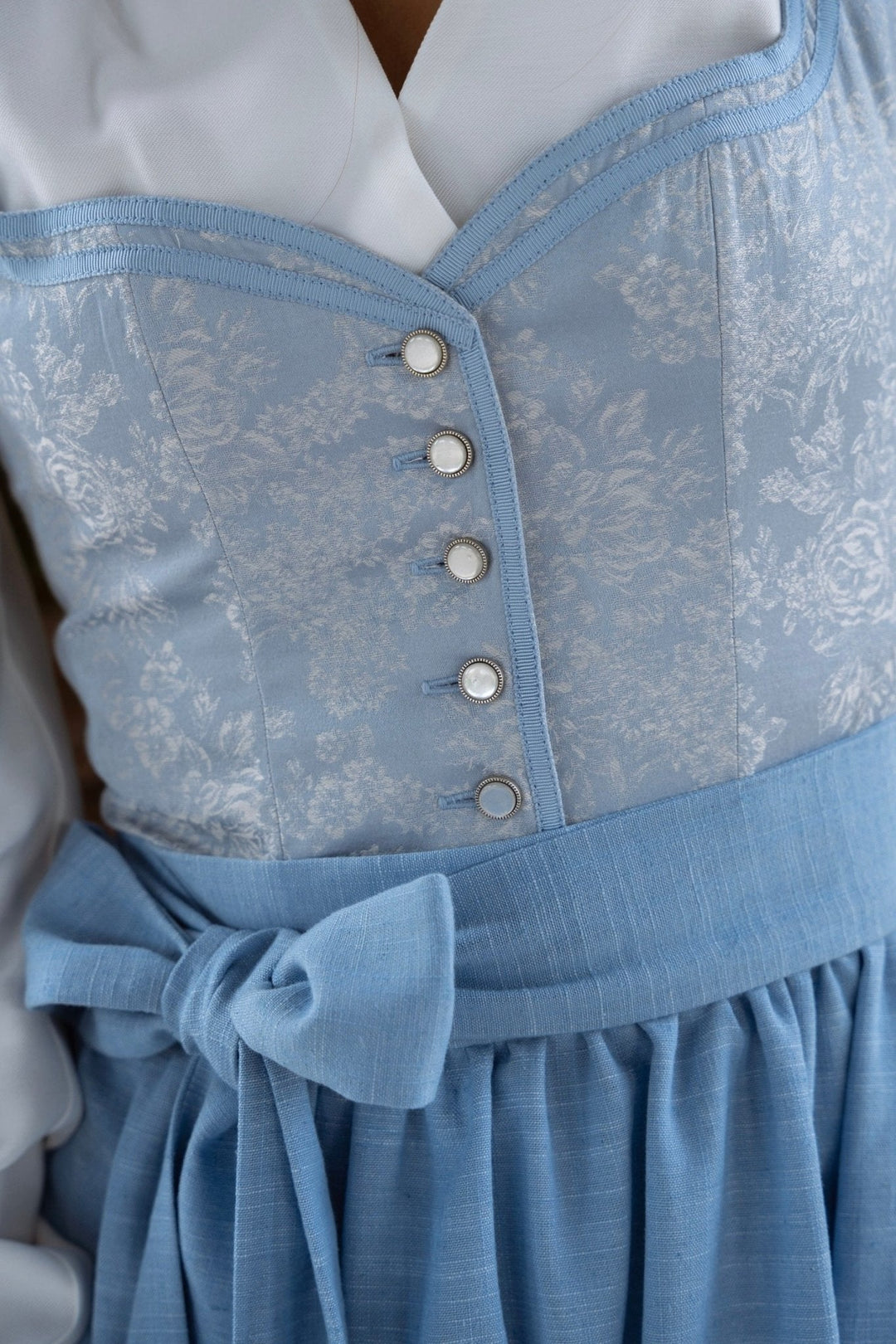 Részlet a világoskék, virágmintás dirndl kötényéről és mellrészéről, masnival és gyöngyházszerű gombokkal.
