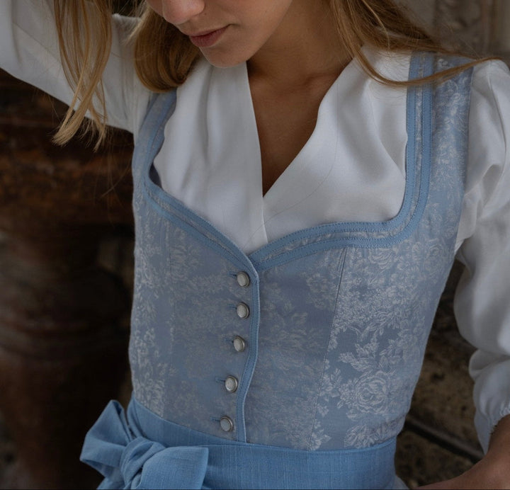 Oldalnézetű világoskék, virágmintás dirndl fehér blúz fölött, gombsorral és szalagszegéllyel.