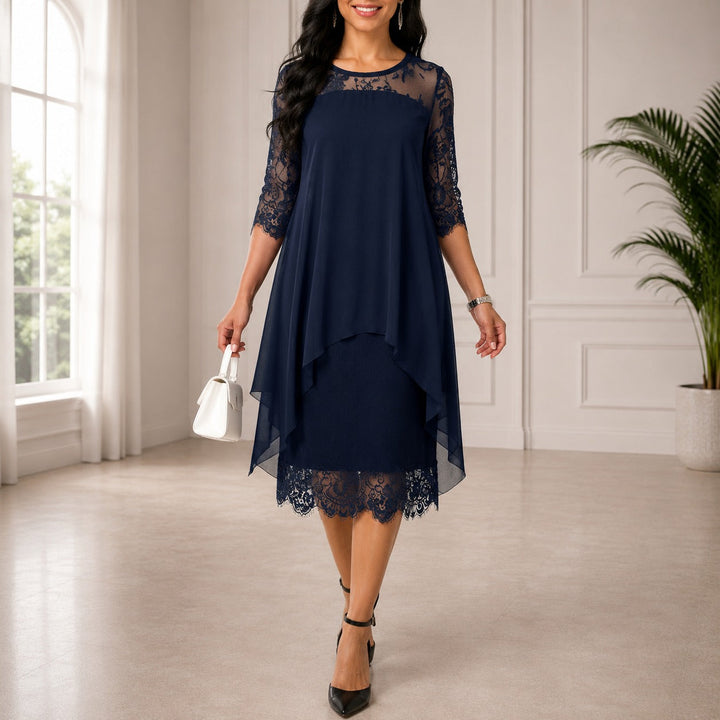 Sötétkék, csipkebetétes ujjú, áttetsző sifon fedőrétegű midi ruha, amelyet fehér kézitáskával és fekete magas sarkú cipővel viselő modell mutat be.