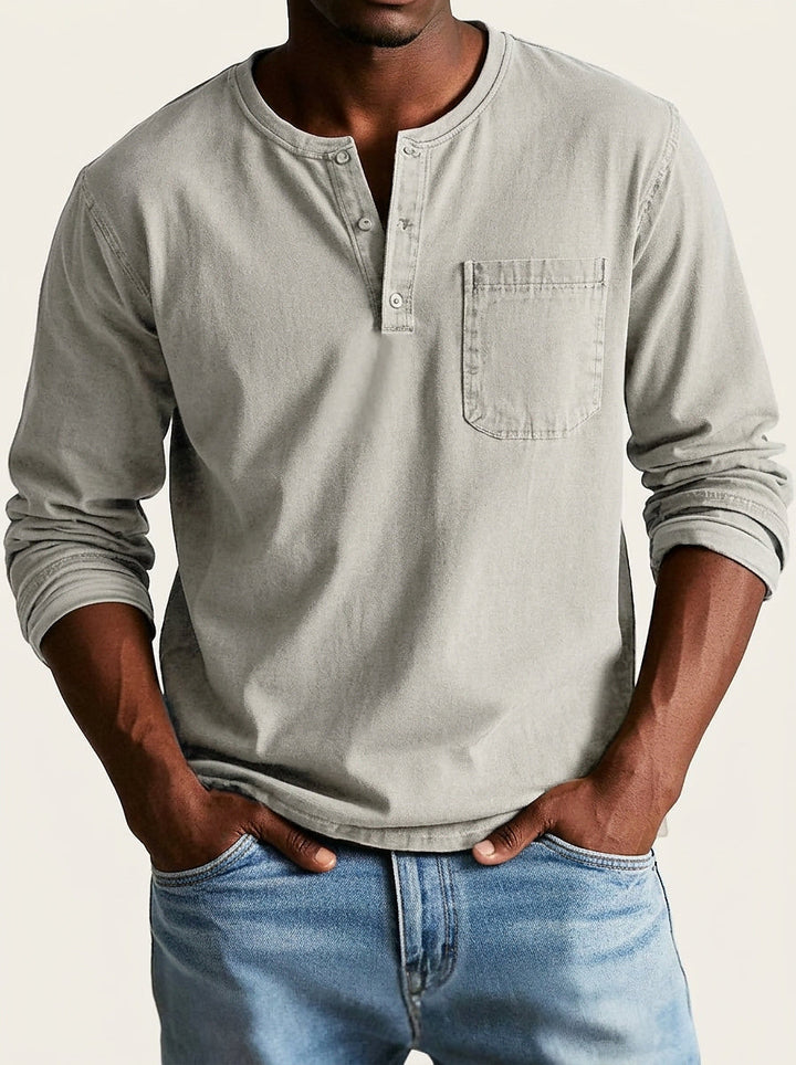 Világos szürkésbézs férfi Henley felső elölről, modellen, farmerrel; a gombos nyakkivágás és a mellzseb jól kivehető.