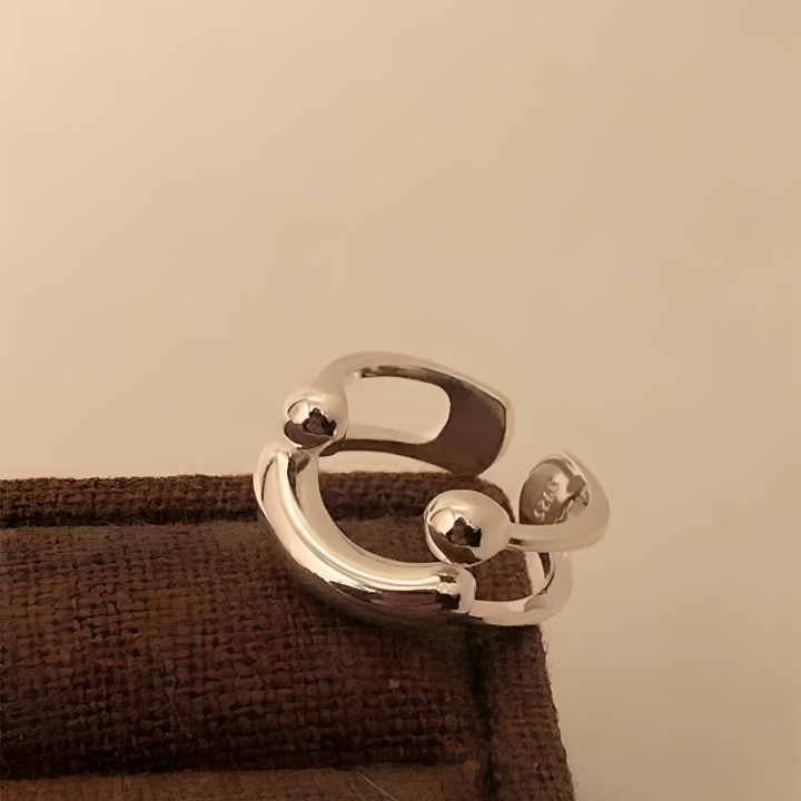 A gyűrű oldalról és kissé felülről, barna textilfelületen, a fényes gömbök és a nyitott, áttört forma részletével.